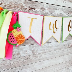 May include: A white banner with gold lettering that reads "TWO" with pink, yellow, and green borders. The banner is decorated with paper cutouts of watermelon, pineapple, and orange slices. The banner is hanging on a white wooden background with colorful ribbons.