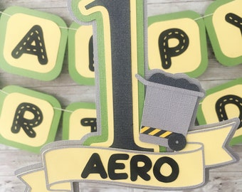 May include: A green, yellow, and gray construction-themed cake topper with a large number one, a small dump truck, and a banner that says "AERO".