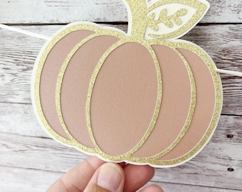 May include: A decorative pumpkin-shaped banner element. The pumpkin is a muted peach color with gold glitter accents outlining the shape and on the stem and leaf. The banner is held by a hand against a light wood background.