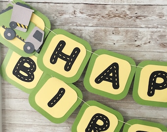 May include: A green and yellow banner with the words "Happy Birthday" in black letters. Each letter is outlined in white and has a road design inside. A small toy dump truck is attached to the banner.