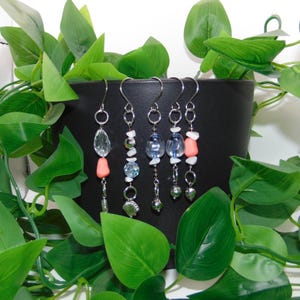 May include: Four pairs of dangle earrings with silver hooks. Each earring features a combination of clear, blue, and coral-colored beads, along with small silver charms. The earrings are displayed against a black background with green foliage.