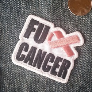 May include: White patch with black text that reads "FU* CANCER" and a pink ribbon. The patch is sewn onto a blue denim background.