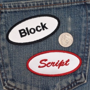 May include: Two white oval patches sewn onto a blue denim background. The top patch has a black border and the text "Block" in black. The bottom patch has a red border and the text "Script" in red.