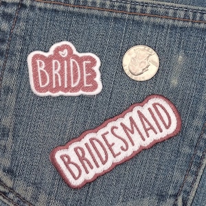 May include: Two pink fabric patches sewn onto a blue denim background. The patches are embroidered with the words "Bride" and "Bridesmaid".