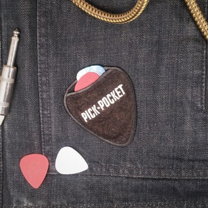 May include: A brown leather guitar pick holder with the text "PICK-POCKET" embroidered on it. The holder is on a dark blue denim background with three guitar picks inside.