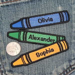 May include: Three iron-on patches shaped like crayons with black outlines. The crayons are blue, green, and orange. The blue crayon says "Olivia", the green crayon says "Alexander", and the orange crayon says "Sophia".