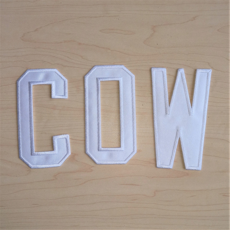 Peut inclure: Trois lettres blanches brod&eacute;es en tissu &eacute;pellent le mot "COW". Les lettres sont soulign&eacute;es d'une bordure blanche et sont pos&eacute;es sur un fond clair en grain de bois. Les lettres sont probablement des &eacute;cussons ou des appliqu&eacute;s.