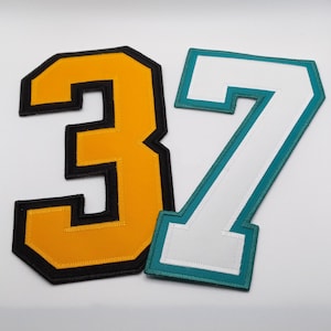 May include: Two felt numbers, a gold 3 and a white 7 with a teal border, are arranged on a white background.