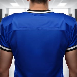 May include: A royal blue football jersey with white and black trim on the collar and sleeves. The jersey has a rectangular patch on the upper back. The image is taken from behind, showing the back of the jersey.