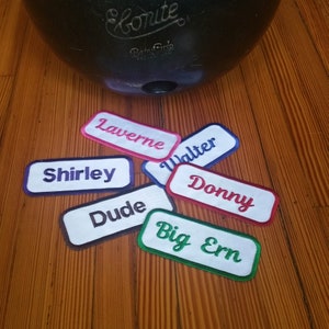 May include: A collection of six colorful embroidered iron-on name patches. The patches are rectangular and feature the names "Shirley", "Laverne", "Walter", "Donny", "Dude", and "Big Ern".