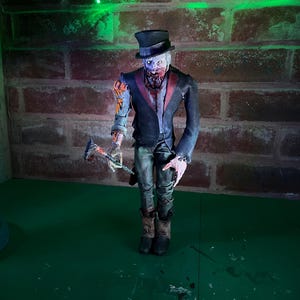 Op de afbeelding: Handgemaakte zombiefiguur met een zwarte hoge hoed, een donkerblauwe jas met rode revers en een zwarte broek. De figuur houdt een bloedige bijl vast en heeft bloot vlees. Achtergrond: bakstenen muur en groen oppervlak.
