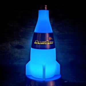 May include: A blue, rocket-shaped lamp with the word "Quantum" in yellow and a small graphic. The lamp has a blue glow and a dark base. The lamp is plugged into a power outlet.