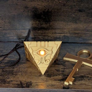 May include: A gold-colored, triangular pendant with an illuminated eye design, a matching key, and a leather cord. The pendant is a pyramid shape with intricate details. The key is a stylized ankh shape.