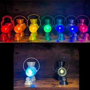 May include: A collection of nine lantern-shaped lamps in various colors including red, orange, yellow, green, light blue, dark blue, purple, white, and black. Each lantern features a circular handle and a glowing light source at its center.