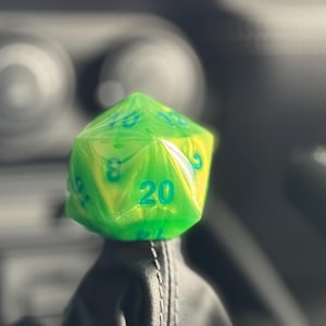 Pode incluir: Um dado de 20 faces verde e azul com números em cada lado. O dado está sentado no topo de uma alavanca de câmbio preta em um carro.