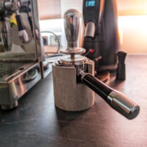 Könnte beinhalten: Ein grauer Betonständer mit einem silbernen Siebträger und Tamper. Der Ständer ist so konzipiert, dass er den Siebträger und den Tamper an Ort und Stelle hält.