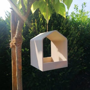 Op de afbeelding: Een lichtgekleurde vogelvoederbak met een glad oppervlak in de vorm van een eenvoudig huisje met een puntdak en een open voorkant. De structuur hangt aan een dun touwtje aan een boomtak en heeft een platte bodem voor voer en een holle binnenkant.