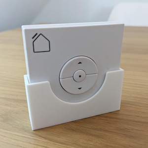 Op de afbeelding: Een witte afstandsbediening voor een slimme woning met een cirkelvormig bedieningspaneel met pijlen omhoog en omlaag en een kleine stip in het midden. De afstandsbediening zit in een witte houder en heeft een huisicoon. Op een houten oppervlak.