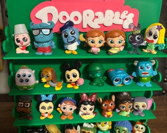 Puede incluir: Estantería verde con varios niveles que contiene una colección de figuras de Disney Doorables. La estantería tiene un letrero rosa con el logotipo de Disney Doorables. Las figuras son coloridas y presentan varios personajes de Disney.
