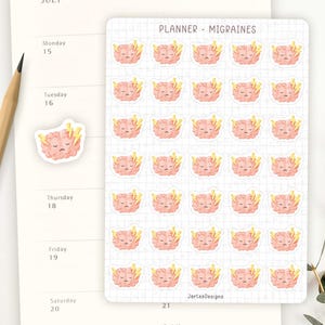 May include: A sheet of planner stickers labelled "PLANNER - MIGRAINES" featuring pink brain illustrations with yellow lightning bolts. A single sticker and a wooden pencil rest on a calendar page for July.