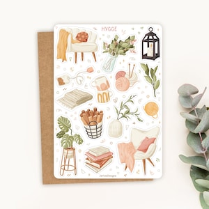 Op de afbeelding: Een stickerblad met een gezellig thema, met illustraties van een stoel, deken, boeken, planten en andere items. Het woord "HYGGE" staat bovenaan. Het stickerblad ligt op een bruine kaart.