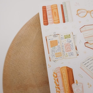 May include: A sticker sheet featuring illustrations of books, an open journal with handwritten notes, a stack of books, a pair of light-colored glasses, and a candle. The designs are rendered in a soft color palette of orange, cream, and muted green.