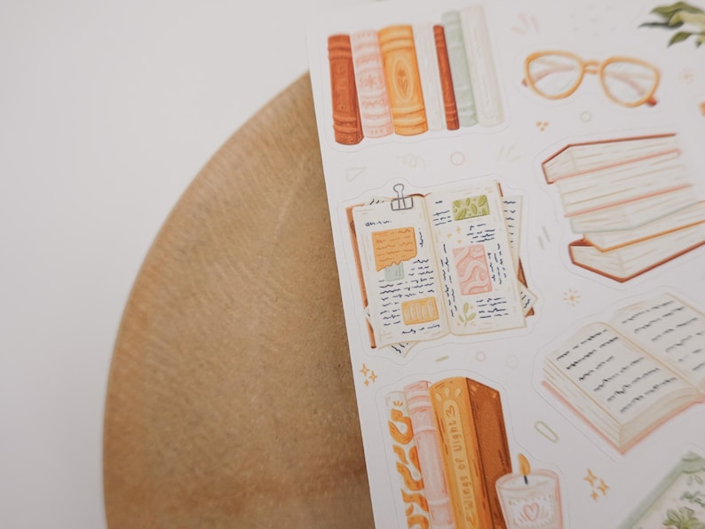 May include: A sticker sheet featuring illustrations of books, an open journal with handwritten notes, a stack of books, a pair of light-colored glasses, and a candle. The designs are rendered in a soft color palette of orange, cream, and muted green.