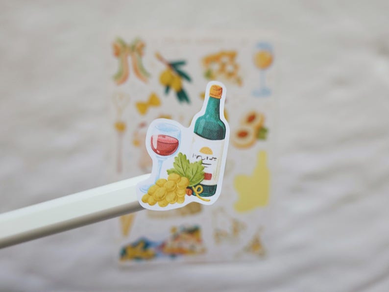 May include: A sticker featuring a wine bottle, a glass of red wine, and a bunch of grapes. The bottle is green with a yellow label. The glass is filled with red wine. The grapes are yellow. The sticker is held by a white stylus.