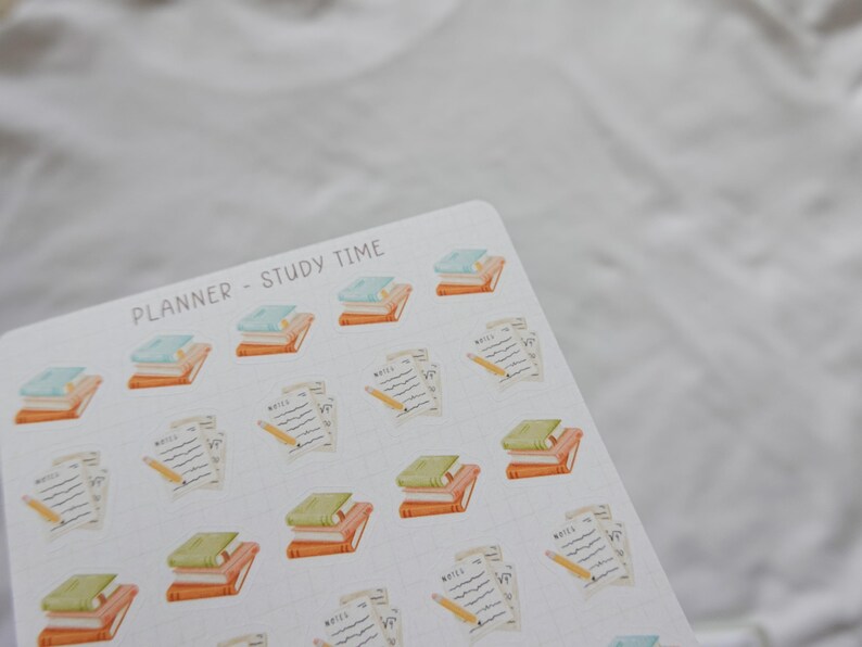 May include: A sheet of planner stickers with a study theme. The stickers feature stacks of books in orange, green, and blue, along with pencils and notepaper. The text "PLANNER - STUDY TIME" is printed at the top.
