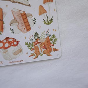 May include: A sticker sheet featuring various illustrations including a brown journal with a mushroom, a stack of books labeled Mycology, several tan and red-capped mushrooms, a tree stump with foliage, and a mushroom-shaped house with the text JorteaDesigns.