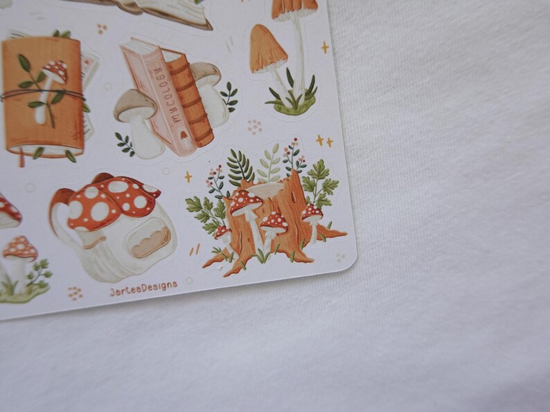 May include: A sticker sheet featuring various illustrations including a brown journal with a mushroom, a stack of books labeled Mycology, several tan and red-capped mushrooms, a tree stump with foliage, and a mushroom-shaped house with the text JorteaDesigns.