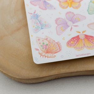 May include: A sticker sheet featuring various watercolor-style moths in pastel shades of pink, yellow, purple, and green. The moths display intricate wing patterns with small crescent moon motifs and floral accents. The text JarteaDesigns is visible.