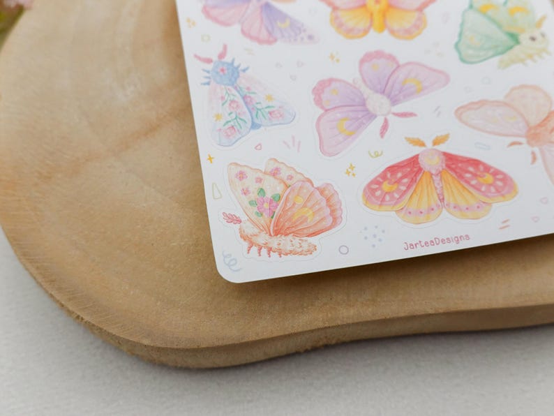 May include: A sticker sheet featuring various watercolor-style moths in pastel shades of pink, yellow, purple, and green. The moths display intricate wing patterns with small crescent moon motifs and floral accents. The text JarteaDesigns is visible.