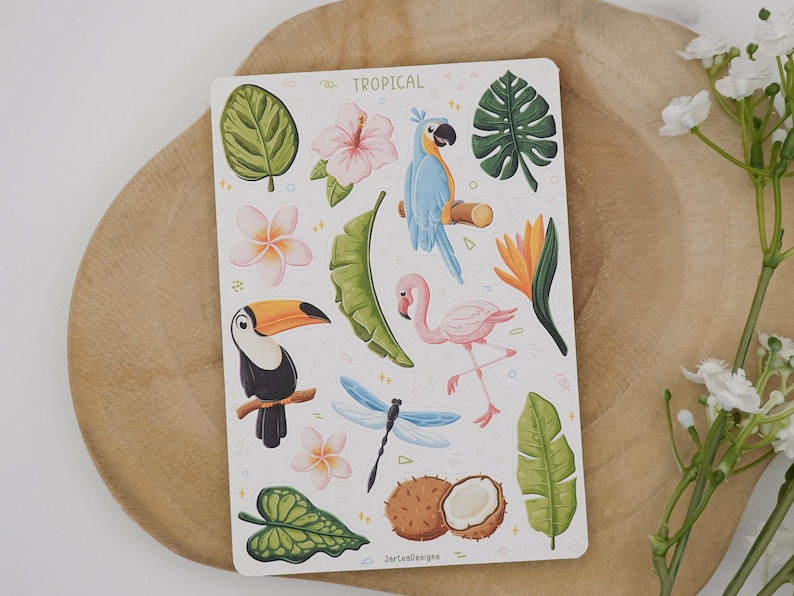 May include: A rectangular sticker sheet titled TROPICAL featuring illustrations of a blue parrot, a pink flamingo, a toucan, a dragonfly, a coconut, various tropical leaves, and flowers including hibiscus and plumeria on a white background.