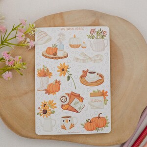 May include: A sticker sheet with autumn-themed illustrations, including pumpkins, sunflowers, and coffee cups. The text "AUTUMN VIBES" is at the top. The sheet is on a wooden surface with pink and red pencils.