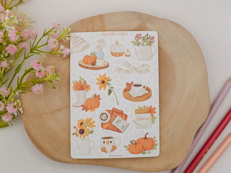 May include: A sticker sheet with autumn-themed illustrations, including pumpkins, sunflowers, and coffee cups. The text "AUTUMN VIBES" is at the top. The sheet is on a wooden surface with pink and red pencils.