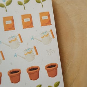 May include: A sheet of stickers with gardening illustrations. The stickers depict orange seed packets, white watering cans with orange handles, brown flower pots, and green plant sprouts. The stickers are on a white background.