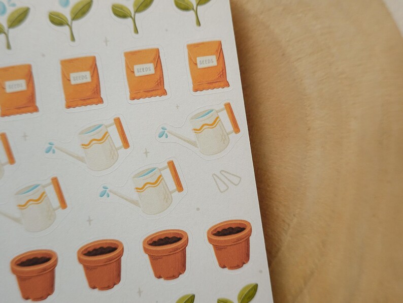 May include: A sheet of stickers with gardening illustrations. The stickers depict orange seed packets, white watering cans with orange handles, brown flower pots, and green plant sprouts. The stickers are on a white background.