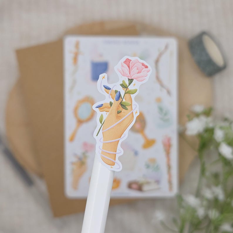 K&ouml;nnte beinhalten: Ein gestanzter Aufkleber mit einer stilisierten Illustration einer Hand mit blau lackierten Fingern&auml;geln, die eine rosa Rose h&auml;lt. Die Hand ist von einer d&uuml;nnen, geschwungenen wei&szlig;en Linie umwickelt und auf einem wei&szlig;en Stift vor unscharfem Hintergrund.