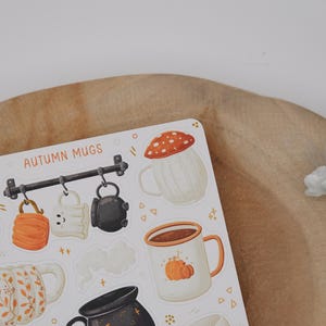 May include: A sticker sheet with illustrations of autumn mugs. Designs include a mushroom-topped mug, a pumpkin-decorated mug, and mugs with fall leaf patterns. The words "AUTUMN MUGS" are printed at the top of the sheet.