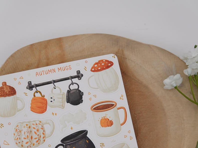 May include: A sticker sheet with illustrations of autumn mugs. Designs include a mushroom-topped mug, a pumpkin-decorated mug, and mugs with fall leaf patterns. The words "AUTUMN MUGS" are printed at the top of the sheet.