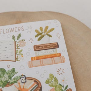 Peut inclure: Une planche d'autocollants illustrant des livres, des plantes en pot et des lunettes de lecture. Le texte "BOOKS & FLOWERS" est imprim&eacute; en haut. Les motifs incluent une pile de quatre livres, dont un marqu&eacute; "LOVE", et divers feuillages verts.
