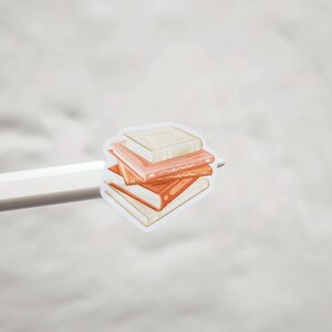 Op de afbeelding: Een sticker met een stapel boeken in oranje en cr&egrave;metinten. De boeken zijn op elkaar gestapeld, waarbij het bovenste boek lichter is dan de andere. De sticker wordt vastgehouden door een witte potlood.