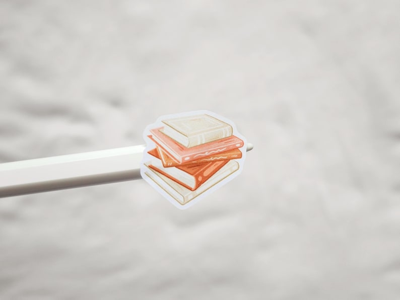 Op de afbeelding: Een sticker met een stapel boeken in oranje en cr&egrave;metinten. De boeken zijn op elkaar gestapeld, waarbij het bovenste boek lichter is dan de andere. De sticker wordt vastgehouden door een witte potlood.