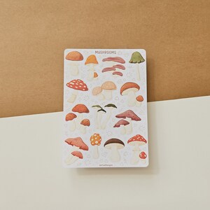 K&ouml;nnte beinhalten: Eine Bogen mit Aufklebern, die verschiedene illustrierte Pilze in Rot-, Orange-, Braun- und Gr&uuml;nt&ouml;nen zeigen. Die Aufkleber befinden sich auf einem wei&szlig;en Hintergrund mit dem Wort "Mushrooms" oben.