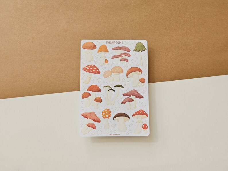 K&ouml;nnte beinhalten: Eine Bogen mit Aufklebern, die verschiedene illustrierte Pilze in Rot-, Orange-, Braun- und Gr&uuml;nt&ouml;nen zeigen. Die Aufkleber befinden sich auf einem wei&szlig;en Hintergrund mit dem Wort "Mushrooms" oben.