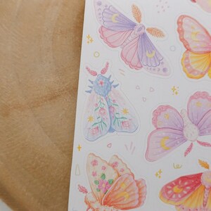 May include: A sticker sheet featuring various moth illustrations in pastel shades of pink, purple, yellow, and orange. The moths are decorated with celestial symbols like crescent moons and stars, along with small floral patterns and tiny geometric shapes.