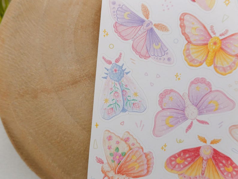 May include: A sticker sheet featuring various moth illustrations in pastel shades of pink, purple, yellow, and orange. The moths are decorated with celestial symbols like crescent moons and stars, along with small floral patterns and tiny geometric shapes.