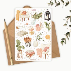 Op de afbeelding: Een stickervel met diverse illustraties met een gezellig thema, waaronder een kleine fauteuil, garen, boeken en een lantaarn. Het woord "HYGGE" staat bovenaan. Een potlood en een envelop staan op de achtergrond.