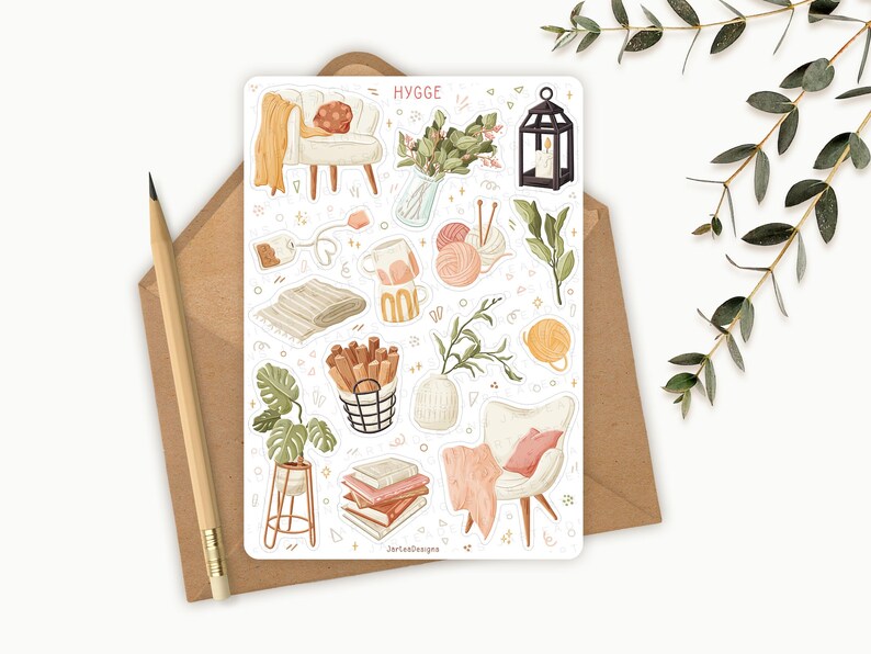 Op de afbeelding: Een stickervel met diverse illustraties met een gezellig thema, waaronder een kleine fauteuil, garen, boeken en een lantaarn. Het woord "HYGGE" staat bovenaan. Een potlood en een envelop staan op de achtergrond.